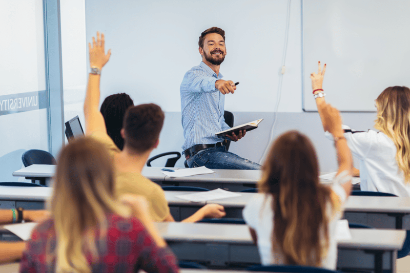 Male teacher in blue shirt and jeans, pointing at a student who has their hand up